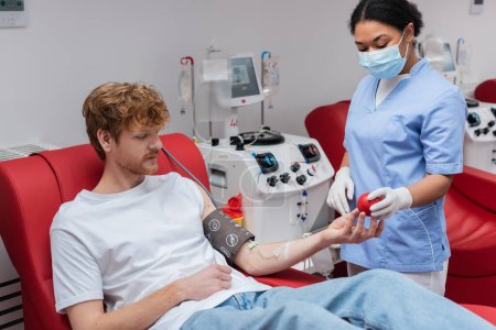 Gesundheitshelfer mit medizinischer Maske und Latex-Handschuhen gibt einem rothaarigen Mann in Blutdruckmanschette Gummiball, der in der Nähe einer Transfusionsmaschine im Krankenhaus auf einem Behandlungsstuhl sitzt
