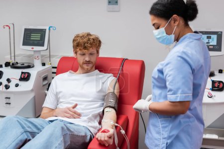 rothaarige Freiwillige mit Transfusionsset, das Gummibälle quetscht, während sie auf einem medizinischen Stuhl in der Nähe automatisierter Geräte sitzt und multirassische Krankenschwester in medizinischer Maske im Blutspendezentrum