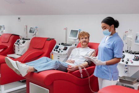 enfermera multirracial en máscara médica y guantes de látex utilizando tableta digital cerca de pelirrojo con transfusión de sangre conjunto sentado en silla ergonómica cómoda al lado de equipo automatizado en la clínica