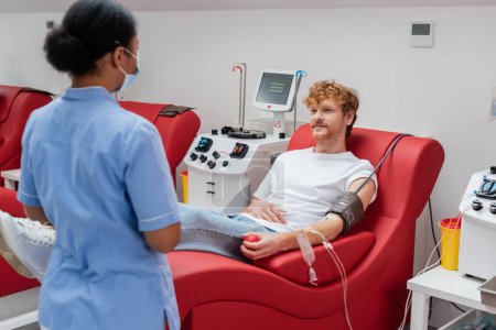 Rotschopf-Mann in Bluttransfusion sitzt auf bequemem Stuhl neben automatisierten Geräten und Krankenschwester in blauer Uniform und medizinischer Maske im Krankenhaus