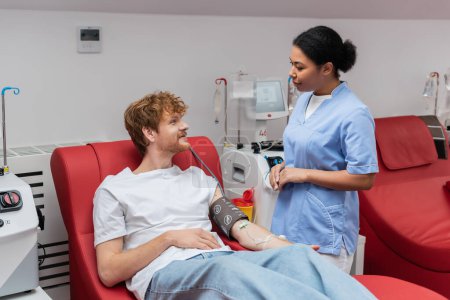 multiracial nurse in blue uniform talking to redhead donor with blood pressure cuff and transfusion set sitting on medical chair near automated equipment in blood donation center