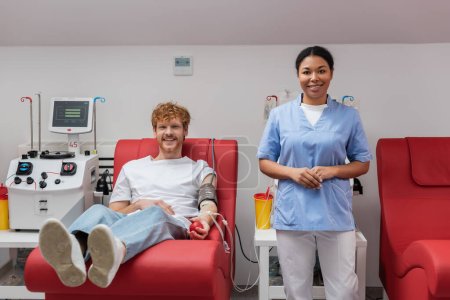 glücklicher rothaariger Mann mit Blutdruckmanschette und Gummiball, der auf einem medizinischen Stuhl in der Nähe einer Krankenschwester sitzt, Transfusionsmaschine und Plastikbechern im Labor und in die Kamera schaut 