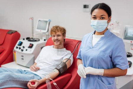 trabajador multirracial de la salud en máscara médica y guantes de látex mirando a la cámara cerca de hombre borroso con transfusión de sangre conjunto sentado en silla ergonómica junto a las máquinas de transfusión en el laboratorio