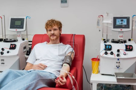 smiling redhead man in transfusion set and blood pressure cuff sitting on medical chair and looking at camera near automated equipment and plastic cup in clinic