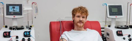 rousse et bénévole positif regardant la caméra tout en étant assis sur une chaise médicale à proximité de machines de transfusion automatisées avec moniteurs dans le centre de don de sang, bannière