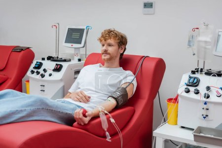 young redhead donor with blood pressure cuff and transfusion set sitting on ergonomic and comfortable medical chair near automated equipment and plastic cups in clinic 