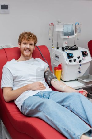 young and cheerful redhead man with transfusion set sitting on ergonomic medical chair and smiling at camera near modern automated equipment and plastic cup