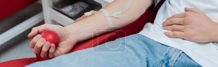 Téléchargez les photos : Vue recadrée du volontaire avec ensemble de transfusion serrant la balle en caoutchouc tout en étant assis sur une chaise médicale confortable dans le centre moderne de don de sang, bannière - en image libre de droit
