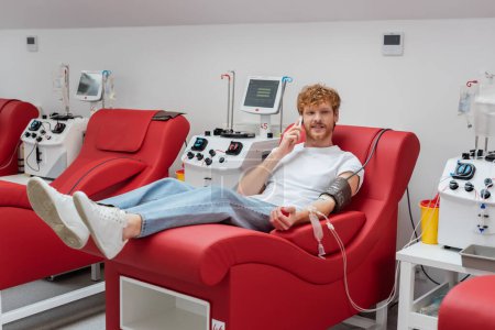 Téléchargez les photos : Jeune homme roux avec un brassard de pression artérielle assis sur une chaise médicale près des machines à transfusion et une tasse en plastique tout en parlant sur un téléphone portable pendant le don de sang à la clinique - en image libre de droit