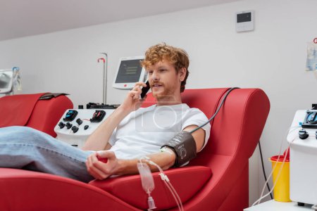 donante pelirrojo con transfusión conjunto sentado en la silla médica y hablando en el teléfono inteligente cerca de equipos automatizados modernos y taza de plástico en el centro de donación de sangre