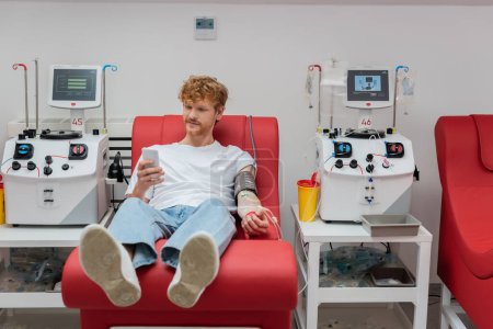Téléchargez les photos : Jeune volontaire rousse assis sur une chaise médicale ergonomique et messagerie sur smartphone pendant la procédure de don de sang près de l'équipement automatisé et tasse en plastique dans la clinique - en image libre de droit