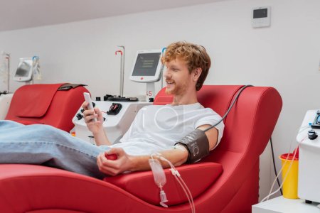 redhead and smiling man browsing internet on mobile phone in medical chair near automated transfusion machines and plastic cup in blood donation center