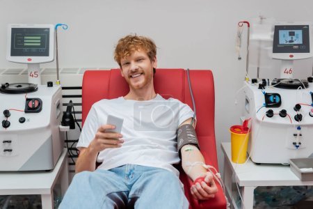homme rousse joyeux avec brassard de pression artérielle et ensemble de transfusion tenant le téléphone portable tout en étant assis sur une chaise médicale près de l'équipement automatisé et tasse en plastique en laboratoire