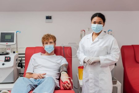 multirassische Arzt in medizinischer Maske in der Nähe Rotschopf Freiwillige mit Blutdruckmanschette und Transfusionsset sitzt auf Medizinerstuhl neben automatischen Geräten und Plastikbecher im Labor