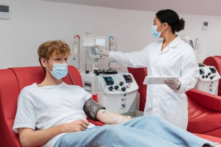 médico multirracial en máscara médica sosteniendo tableta digital y operando la máquina de transfusión cerca de pelirroja voluntario con manguito de presión arterial en el laboratorio