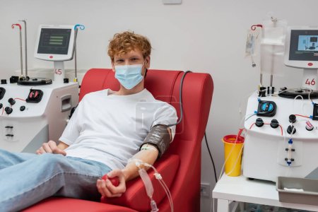 pelirroja voluntaria en máscara médica y manguito de presión arterial mirando a la cámara en silla cómoda cerca de máquinas de transfusión automatizadas en laboratorio moderno