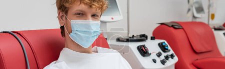 Photo for Young and redhead volunteer in medical mask, with positive eyes expression, donating blood near contemporary transfusion machine and looking at camera in laboratory, banner - Royalty Free Image