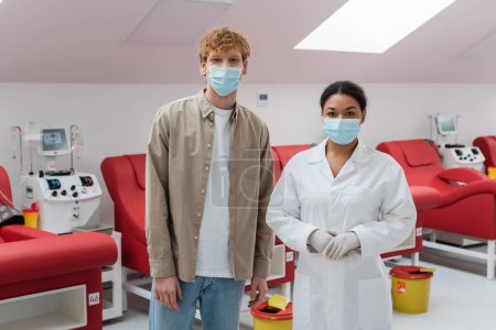 Multirassische Ärztin und rothaarige Freiwillige in medizinischen Masken schauen in die Kamera in der Nähe von Medizinstühlen, Transfusionsmaschinen und Mülleimern im Blutspendezentrum