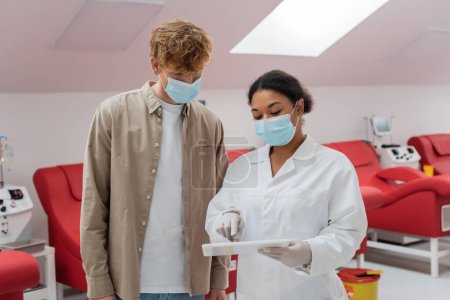 multiracial doctor in medical mask pointing at digital tablet near redhead man and medical chairs with transfusion machines in blood donation center