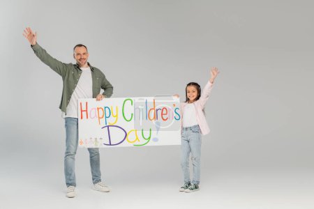 Cheerful father and preteen kid waving hands and holding placard with happy children's day lettering and looking at camera together while standing on grey background