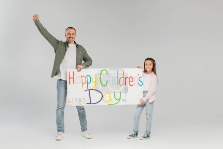 Lächelndes Mädchen in legerer Kleidung mit Plakat mit fröhlichem Kindertag-Schriftzug in der Nähe des Vaters zeigt Ja-Geste und blickt in die Kamera auf grauem Hintergrund