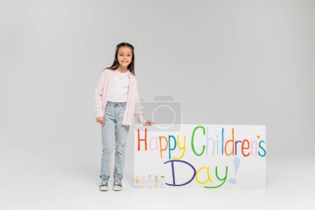 Smiling preteen girl in casual clothes looking at camera while standing near placard with happy children's day lettering during holiday in June on grey background with copy space 