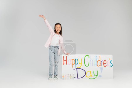 In voller Länge winkt ein fröhliches Mädchen in lässiger Kleidung mit der Hand und schaut in die Kamera neben einem Plakat mit fröhlichem Kindertagsaufdruck auf grauem Hintergrund.