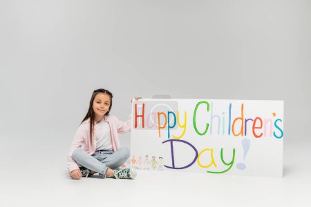 Preteen fille souriante en vêtements décontractés regardant la caméra tout en étant assis près de la pancarte avec le lettrage heureux de la journée des enfants pendant les vacances en Juin sur fond gris