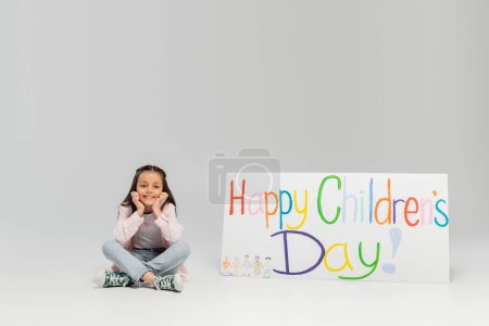 Overjoyed preteen girl in casual clothes sitting and looking at camera near placard with happy children's day lettering during celebration in June on grey background with copy space