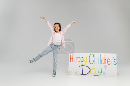 Aufgeregtes und positives Mädchen in lässiger Kleidung, das Spaß hat und auf ein Plakat mit fröhlichem Kindertagsaufdruck während der Feier auf grauem Hintergrund blickt