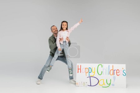 Full length of cheerful father in casual clothes holding preteen daughter on hands near placard with happy children's day lettering during celebration on grey background