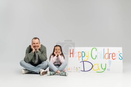 Smiling father and preteen daughter in casual clothes looking at camera while sitting near placard with happy children's day lettering during celebration in June on grey background