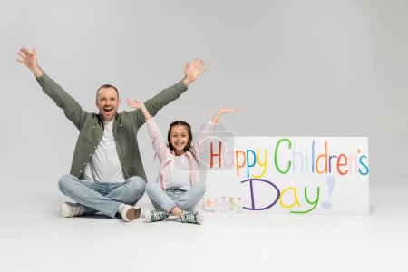 Excited and cheerful man and preteen daughter in casual clothes looking at camera together while sitting near placard with happy children's day lettering on grey background