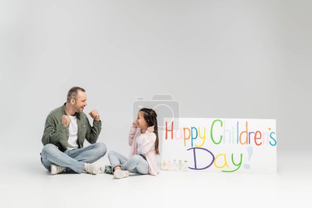 Excited father and daughter in casual clothes looking at each other while sitting near placard with happy children's day lettering during celebration on grey background