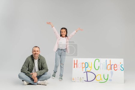 Aufgeregtes junges Mädchen in lässiger Kleidung blickt in die Kamera, während es neben Vater steht und Plakat mit fröhlichem Kindertagsaufdruck im Urlaub auf grauem Hintergrund