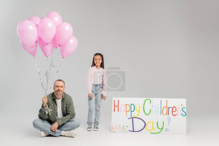 Lächelndes Mädchen in legerer Kleidung, das in die Kamera neben Vater blickt und Luftballons und Plakate mit fröhlichem Kindertagsaufdruck auf grauem Hintergrund während der Feier hält 