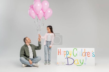 Smiling father giving festive balloons to preteen daughter in casual clothes near placard with happy children's day lettering during event in June on grey background