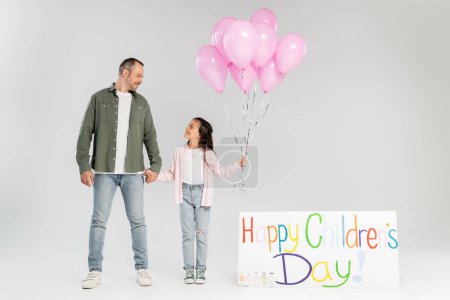 Fröhliches Mädchen in legerer Kleidung mit festlichen Luftballons und Papas Hand, während es bei der Feier im Juni auf grauem Grund neben einem Plakat mit fröhlichem Kindertagsaufdruck steht