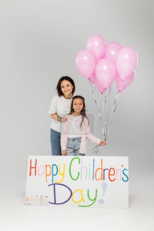 Foto de Sonriente madre tatuada en ropa casual abrazando a la hija preadolescente con globos rosados cerca de la pancarta con letras felices del día de los niños y celebrando en junio sobre fondo gris - Imagen libre de derechos