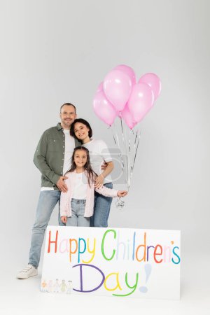 Foto de Los padres sonrientes en ropa casual abrazando a la hija preadolescente con globos rosados cerca de la pancarta con letras felices del día de los niños mientras celebran juntos en junio sobre un fondo gris - Imagen libre de derechos