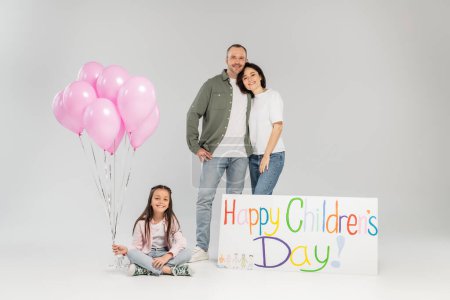 Smiling preteen girl in casual clothes holding festive balloons while sitting near parents hugging and placard with happy children's day lettering on grey background with copy space