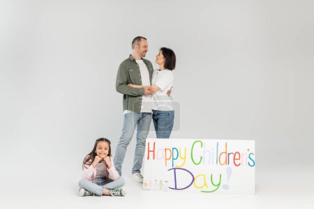 Smiling preteen girl in casual clothes looking at camera while parents hugging and standing beside placard with happy children's day lettering during celebration on grey background