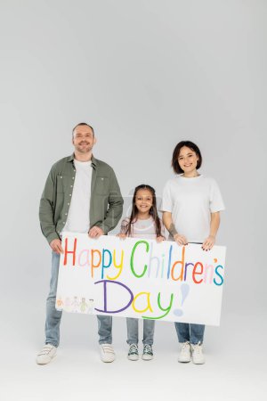 Full length of smiling preteen girl in casual clothes looking at camera while holding placard with happy children's day lettering with parents during event in June on grey background
