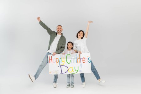 Full length of excited parents in casual clothes looking at camera while holding placard with happy children's day lettering near preteen daughter during holiday in June on grey background