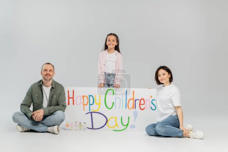 Full length of preteen kid in casual clothes looking at camera while standing near smiling parents and placard with happy children's day lettering during holiday on grey background
