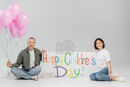 Smiling adult family in casual clothes looking at camera while holding pink festive balloons and placard with happy children's day lettering on grey background with copy space