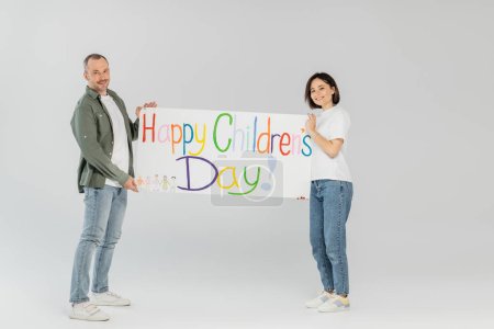 Full length of smiling adult couple in casual clothes looking at camera while holding placard with happy children's day lettering together while standing on grey background