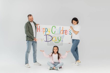 Smiling parents holding placard with happy children's day lettering and looking at preteen daughter pointing with hands during celebration in June on grey background