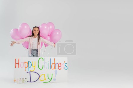 Chica alegre y preadolescente en ropa casual mirando a la cámara cerca de globos festivos rosados y pancarta con letras felices del día de los niños mientras celebra sobre fondo gris