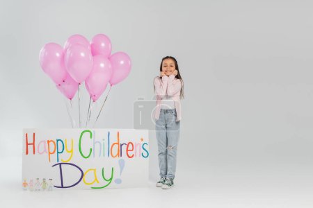 Unbekümmertes Mädchen in lässiger Kleidung, das in die Kamera blickt, während es neben rosa Luftballons und einem Plakat mit fröhlichem Kindertagsaufdruck auf grauem Hintergrund steht